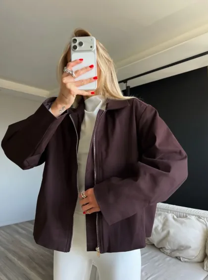 Person takes a mirror selfie wearing an oversized dark brown zip-front jacket, white top, and white pants; red nails and rings visible as they hold a silver iPhone to cover their face.