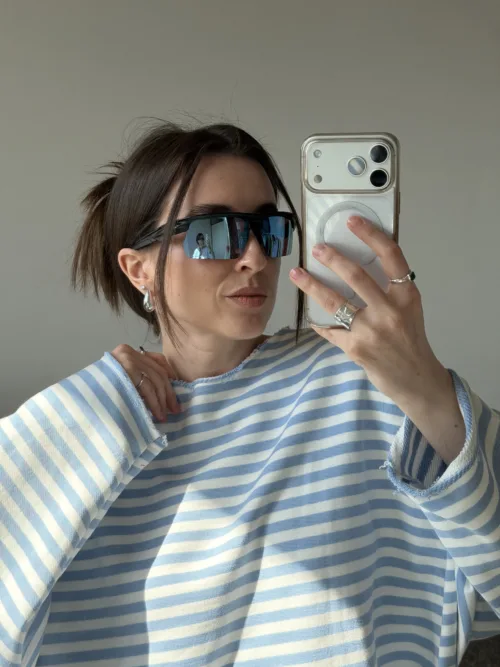 Woman taking a mirror selfie wearing blue-and-white striped top and oversized sunglasses, holding a silver phone with a clear case.