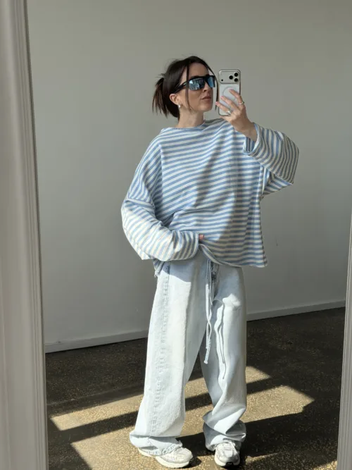 Person taking a mirror selfie in an oversized blue-and-white striped sweater, light jeans, sneakers, and dark sunglasses.