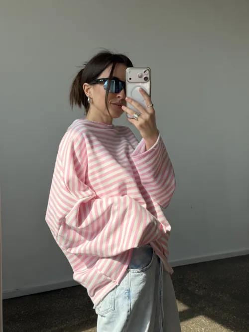 Person taking a mirror selfie in a pink striped sweater and light jeans, wearing dark sunglasses and holding a silver phone.