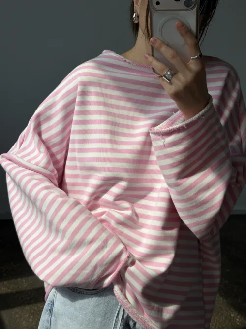 Person in pink and white striped oversized sweater taking a mirror selfie with a phone, jewelry visible (rings and earrings).