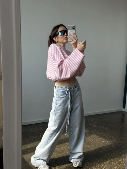 Person taking a mirror selfie wearing a pink and white striped cropped sweater and light baggy jeans, with dark sunglasses indoors.