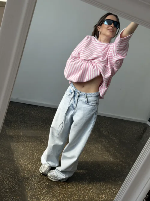Woman in a pink and white striped oversized top tied at the waist, light-wash baggy jeans, sneakers, and blue sunglasses taking a mirror selfie.