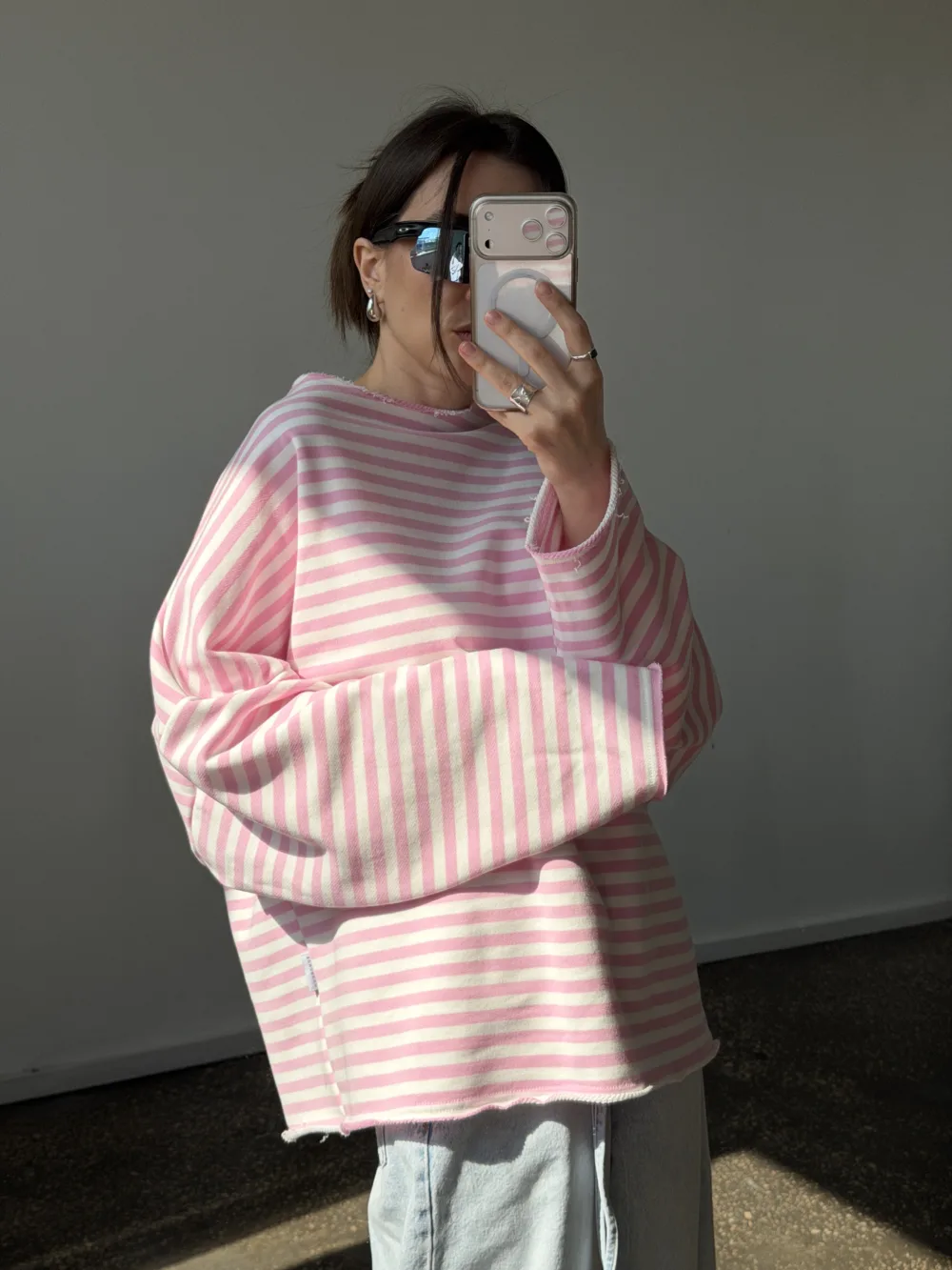 Person in an oversized pink and white striped sweater takes a mirror selfie while wearing dark sunglasses and silver jewelry.