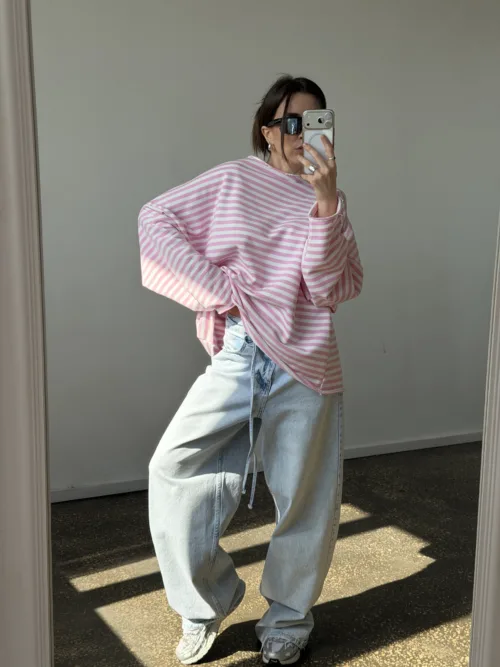 Person taking a mirror selfie in a light-filled room, wearing an oversized pink and white striped sweater, baggy light-wash jeans, white sneakers, and black sunglasses.
