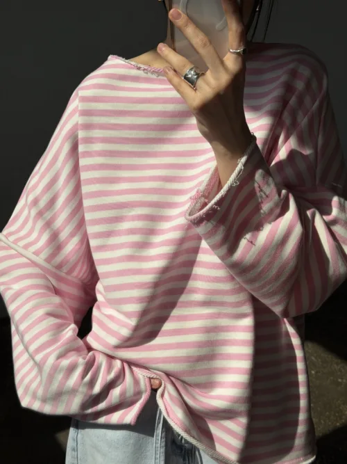 Person wearing a pink and white striped sweater, holding a smartphone up to their face with rings on their fingers and denim jeans visible in the shot.