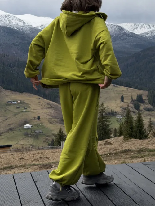 Person wearing a lime-green hoodie and sweatpants stands on a wooden deck overlooking a valley with snow-capped mountains.