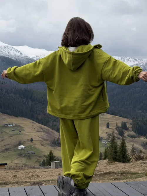 Person in a lime-green hoodie stands on a wooden platform with arms spread, facing snow-capped mountains and a valley below.
