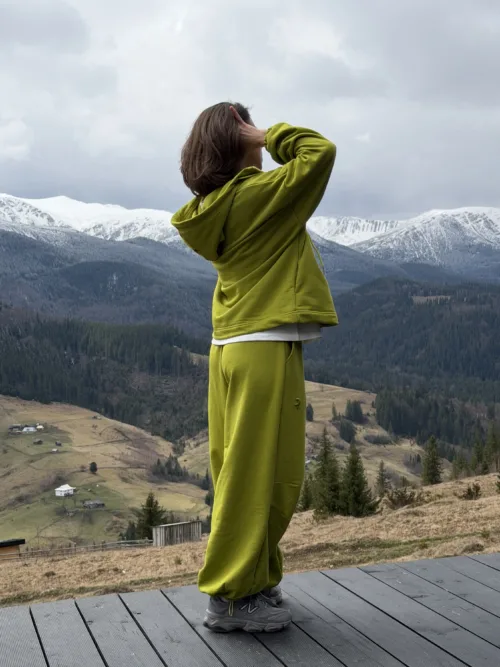 Person wearing a lime-green tracksuit stands on a wooden deck, looking out over snow-capped mountains and a valley.