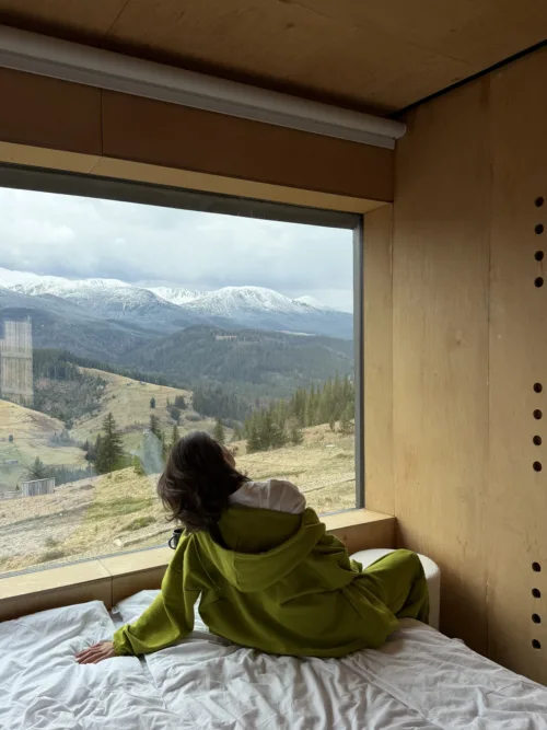 Person in a bright green jacket sits on a bed inside a wooden cabin, looking out at snow-capped mountains through a large window.