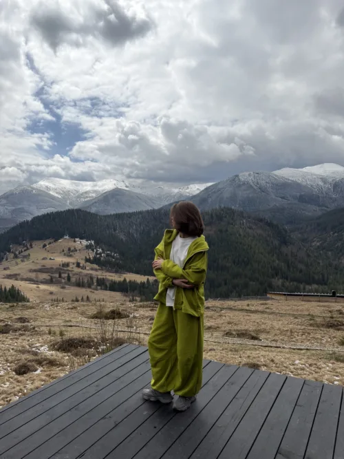 Person wearing a bright lime-green outfit stands on a wooden deck, with snow-capped mountains and cloudy sky in the background