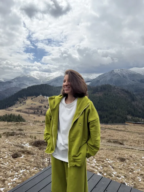 Smiling person in a bright green jacket stands on a wooden platform with snowy mountains and cloudy sky behind.