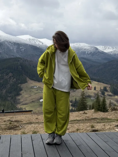 Person in a lime jacket and pants standing on a wooden deck with snow-capped mountains behind him/her.