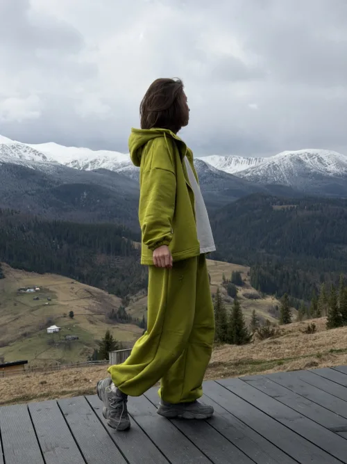 Person in a lime-green tracksuit stands on a wooden deck, gazing at snow-capped mountains.