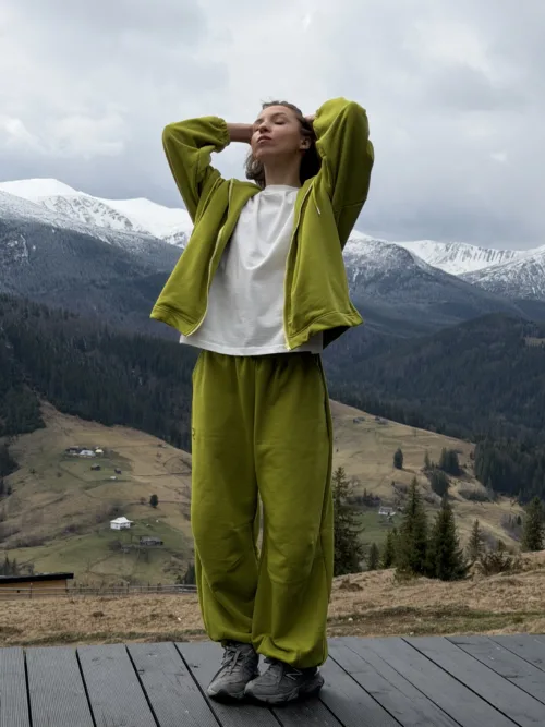 Person standing on a wooden platform in a lime-green tracksuit, arms behind the head, with snow-capped mountains in the background.