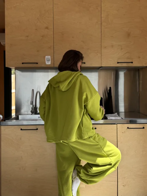 Person wearing a lime green hoodie and pants stands at a stainless kitchen counter, back to the camera, preparing food.