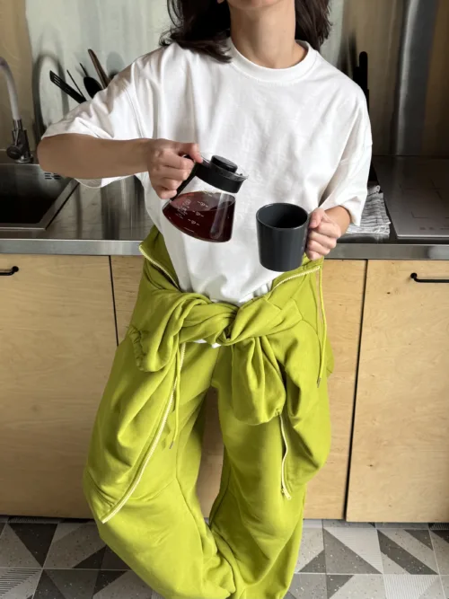 Person in a white t-shirt with a lime-green jacket tied around the waist pours dark coffee from a glass carafe into a black mug in a kitchen.