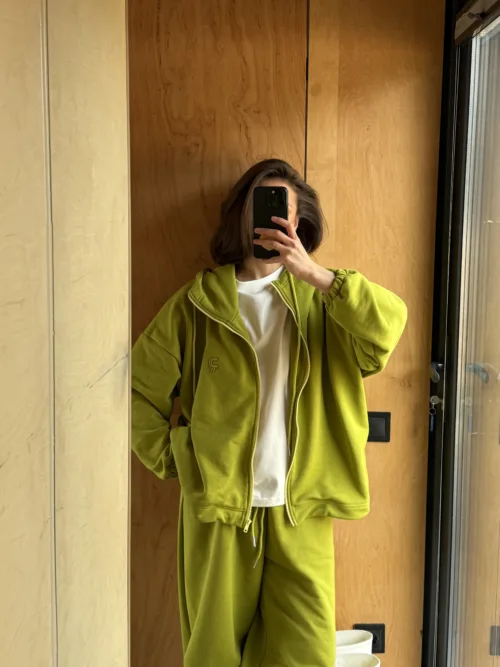 Person wearing a lime-green tracksuit taking a mirror selfie in an elevator with wooden walls and a black phone obscuring their face.
