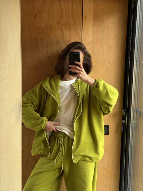 Person wearing a lime-green tracksuit taking a mirror selfie with a black smartphone, face obscured by the device against a wooden wall background.
