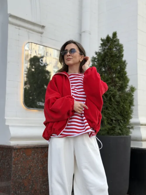 Woman posing in a red bomber jacket over a red-and-white striped shirt and white wide-leg pants, outdoors by a white building with greenery in the background