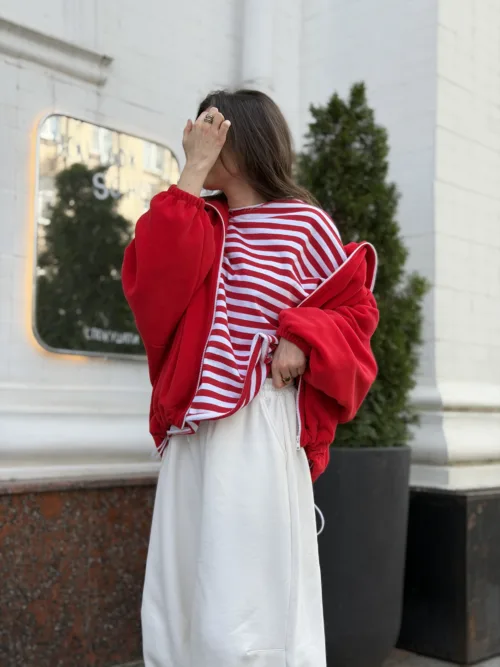 Woman in a red oversized jacket and red-and-white striped top, white wide-leg pants, standing outside a light building, hand raised to cover her face.