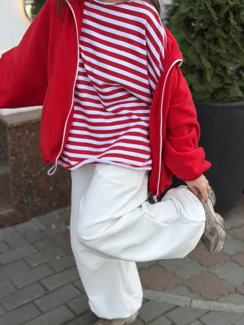 Person in a red jacket and red-and-white striped shirt bends to put on a beige sneaker outdoors on a sidewalk with white pants visible.