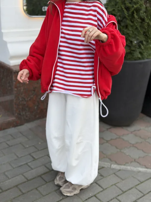 Person wearing a red zip-up jacket over a red-and-white striped shirt, white wide-leg pants, and beige sneakers, standing on a sidewalk outdoor setting.