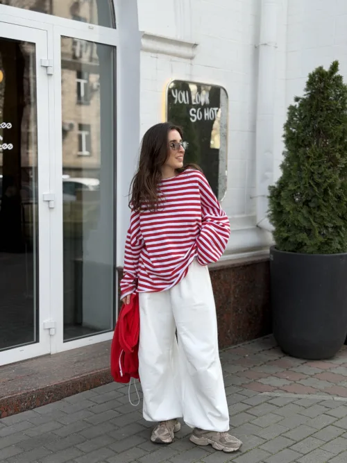 Woman in a red-and-white striped sweater and oversized white pants stands on a sidewalk outside a storefront, holding a red jacket. A sign behind her reads, 'YOU LOOK SO HOT.'