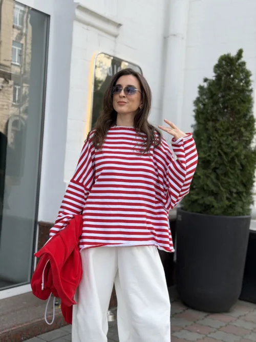 Woman in a red-and-white striped sweater and white pants posing on a city sidewalk, holding a red jacket and wearing sunglasses.