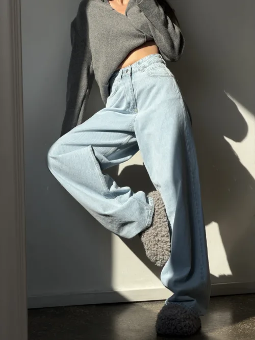 Person wearing a gray oversized sweater and light-wash jeans, posing with one leg raised indoors in soft light.