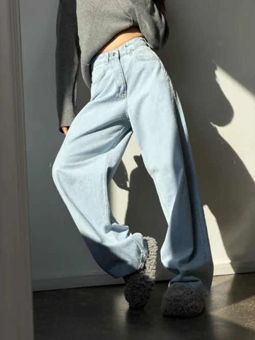 Person wearing light-wash wide-leg jeans and a cropped gray sweater stands in a sunlit doorway, wearing fluffy slippers.