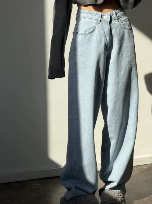 Person standing indoors wearing light-wash wide-leg jeans with a cropped dark sweater and fuzzy gray slippers, bathed in sunlight.