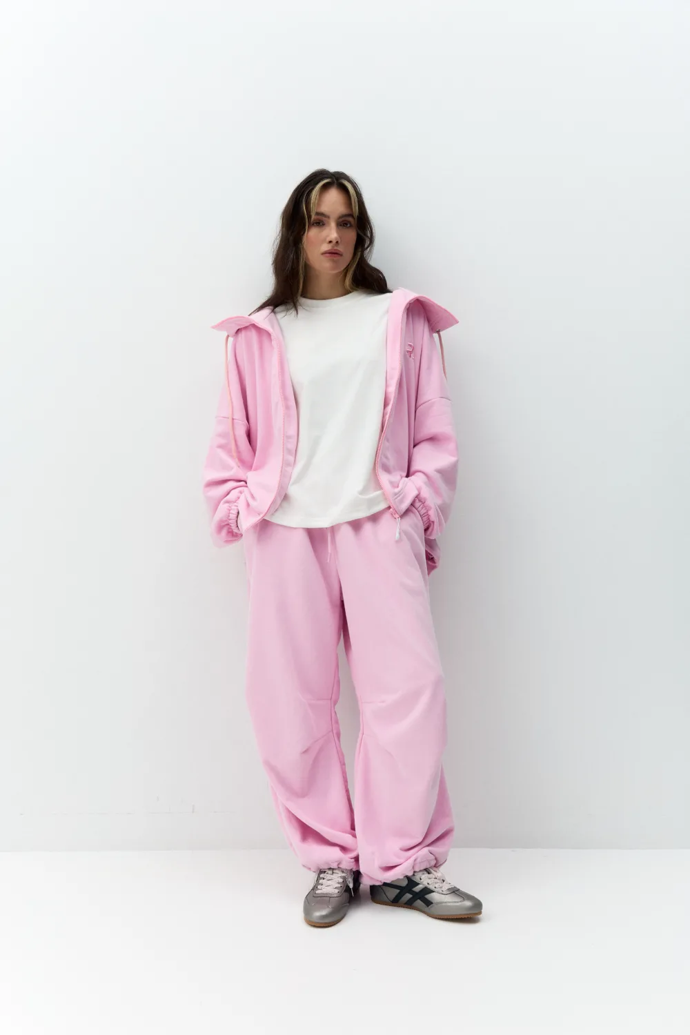 Person in a pastel pink tracksuit with a white T-shirt, standing against a plain white wall, hands in pockets