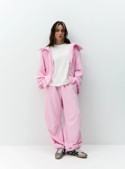 Person in a pastel pink tracksuit with a white T-shirt, standing against a plain white wall, hands in pockets