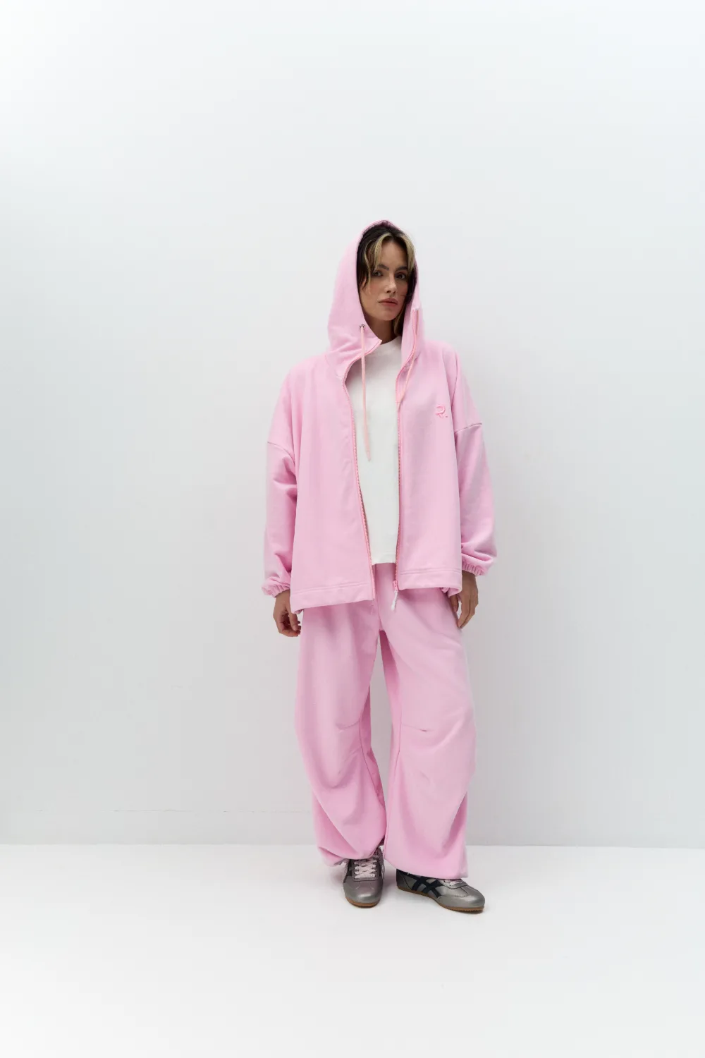 Жіночий сірий oversize костюм з вишивкою та м’якого футеру. Вільний силует і комфортна посадка для повсякденних та relaxed образів. - 1C7A0404_Original Костюм жіночий oversize сірий з вишивкою з футеру - Person wearing a pink hoodie and matching sweatpants, white T-shirt, standing against a plain white wall with hands at sides.