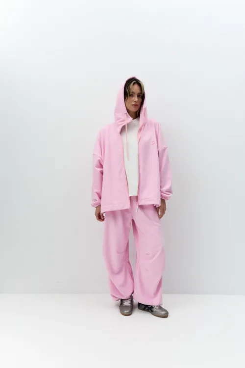 Person wearing a pink hoodie and matching sweatpants, white T-shirt, standing against a plain white wall with hands at sides.