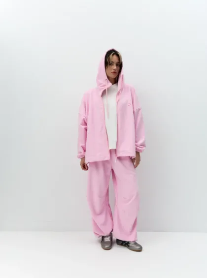 Person wearing a pink hoodie and matching sweatpants, white T-shirt, standing against a plain white wall with hands at sides.