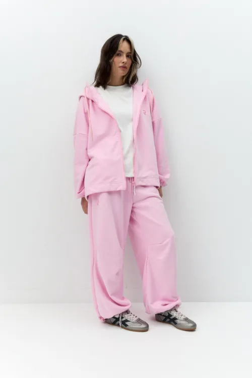 Person wearing a light pink oversized zip-up hoodie and matching pink sweatpants, white tee underneath, standing against a white wall with hands in pockets.