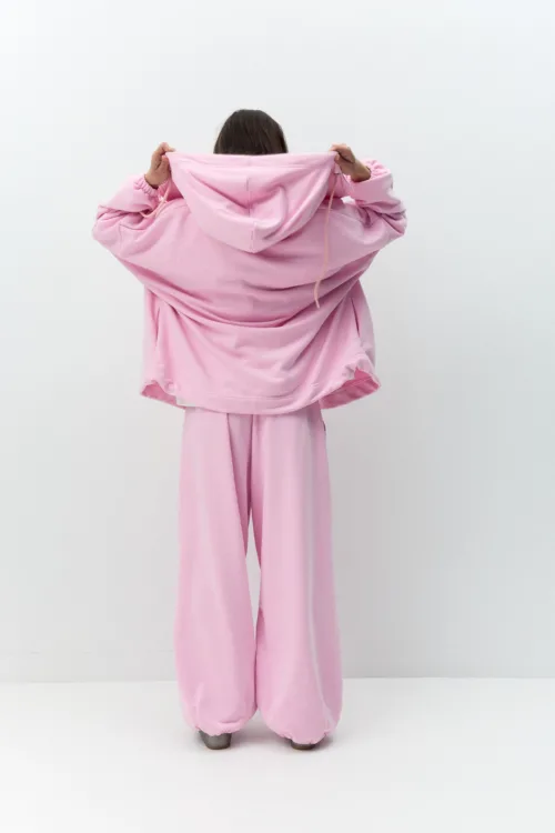 Back view of a person in an oversized pink hoodie and matching pants, holding up the hood with both hands.