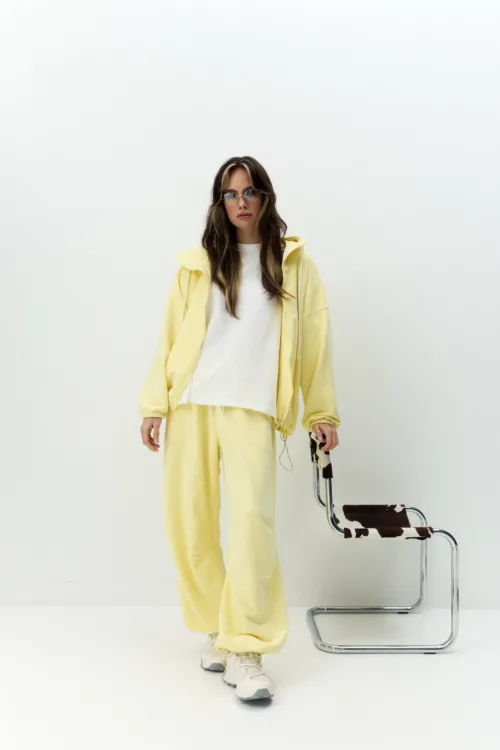 Person in a pale yellow tracksuit and white shirt stands against a white backdrop, wearing sunglasses and sneakers, with a cowhide-patterned chair nearby.