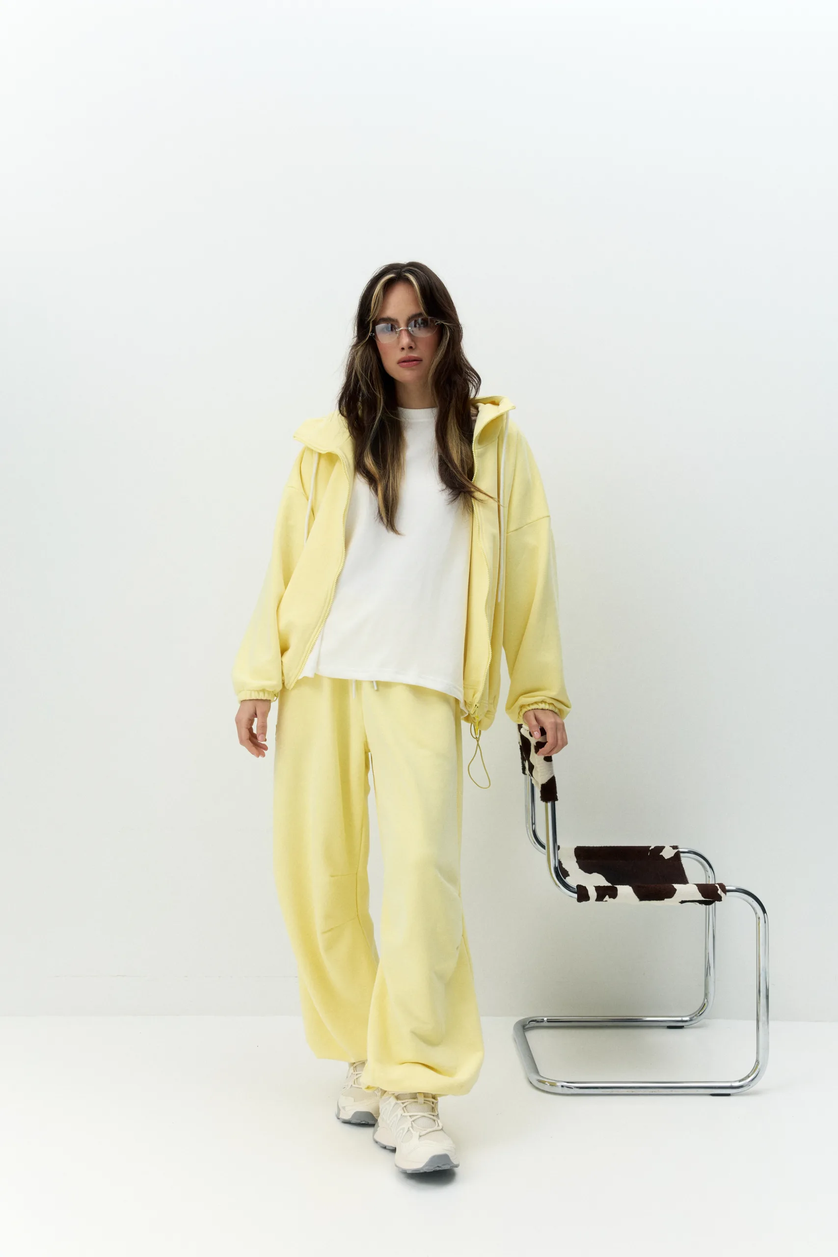 Жіноче oversized худі лимонного кольору з футеру діагональ преміум. Вільний силует, блискавка та м’яка фактура для casual образів. - 1C7A0502_Original Худі жіноче oversized лимонне на блискавці без утеплення - Person in a pale yellow tracksuit and white shirt stands against a white background, wearing sunglasses near a chrome chair with cowhide upholstery