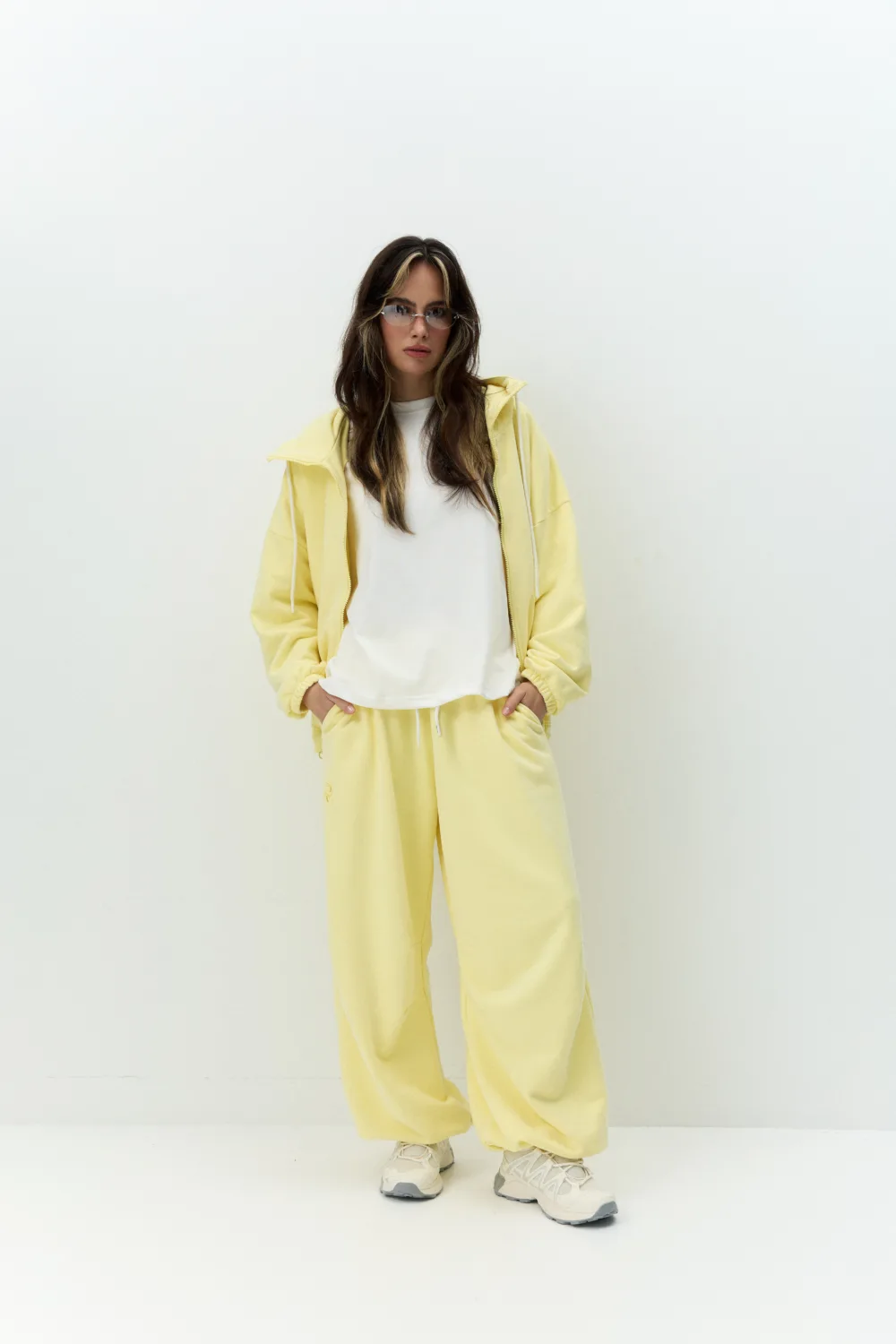 Жіночий сірий oversize костюм з вишивкою та м’якого футеру. Вільний силует і комфортна посадка для повсякденних та relaxed образів. - 1C7A0517_Original Костюм жіночий oversize сірий з вишивкою з футеру - Woman standing against a pale wall wearing a yellow tracksuit with a white shirt, hands in pockets, sneakers visible.