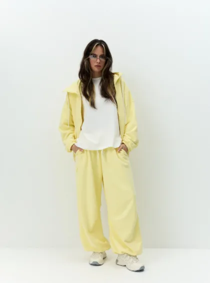 Woman standing against a pale wall wearing a yellow tracksuit with a white shirt, hands in pockets, sneakers visible.
