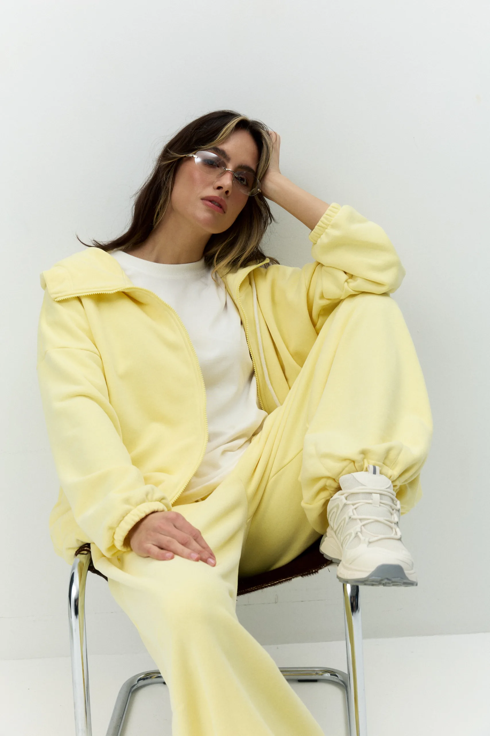 Жіноче oversized худі лимонного кольору з футеру діагональ преміум. Вільний силует, блискавка та м’яка фактура для casual образів. - 1C7A0572_Original Худі жіноче oversized лимонне на блискавці без утеплення - Woman in a pale yellow tracksuit sitting on a chair against a white wall, hand resting on head, wearing sunglasses and white sneakers.