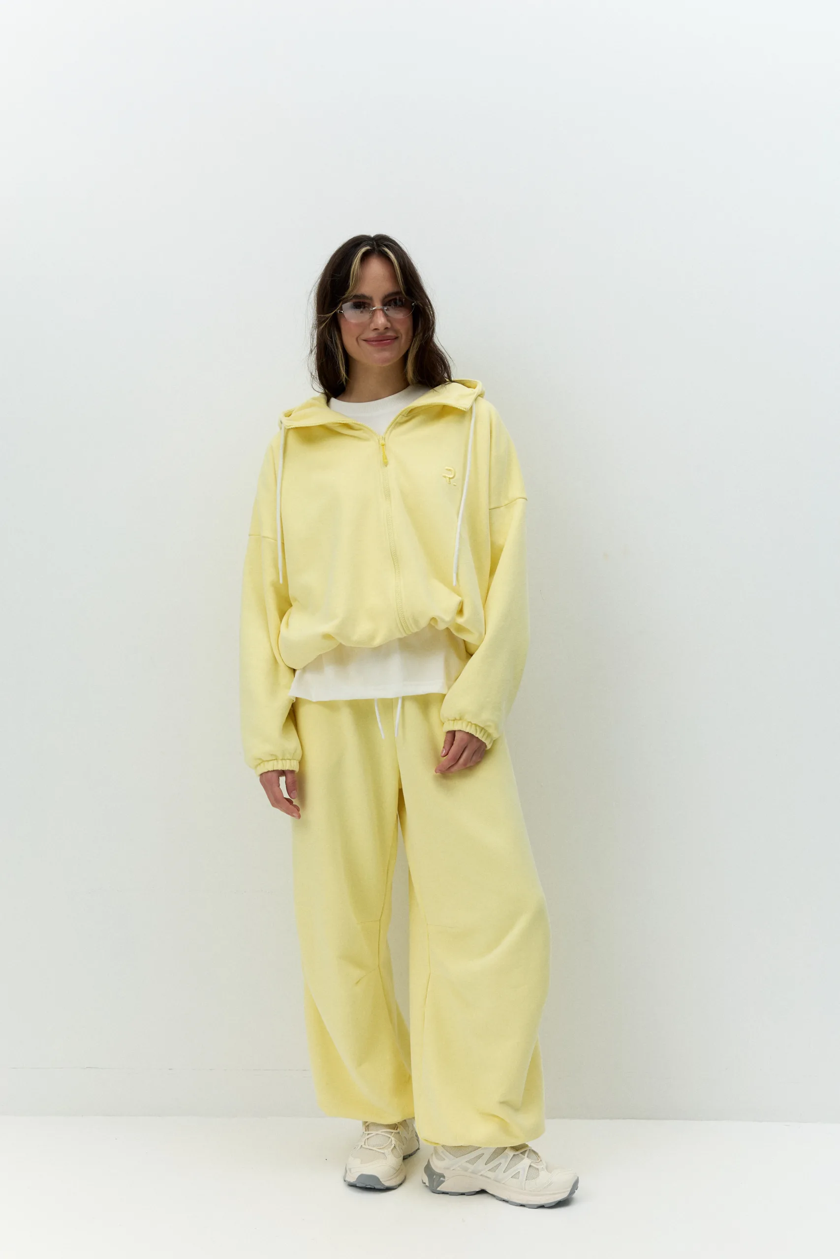 Жіноче oversized худі лимонного кольору з футеру діагональ преміум. Вільний силует, блискавка та м’яка фактура для casual образів. - 1C7A0585_Original Худі жіноче oversized лимонне на блискавці без утеплення - Person wearing a yellow tracksuit with a white shirt, standing against a plain white wall, smiling slightly.