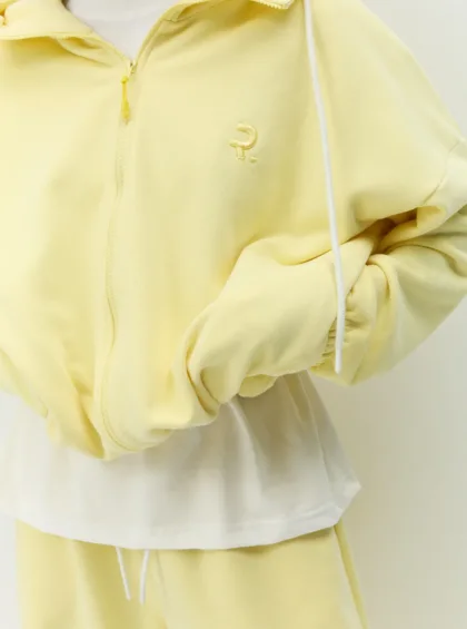 Close-up of a pale yellow zip-up hoodie with white drawstrings and a small embroidered logo on the left chest area.