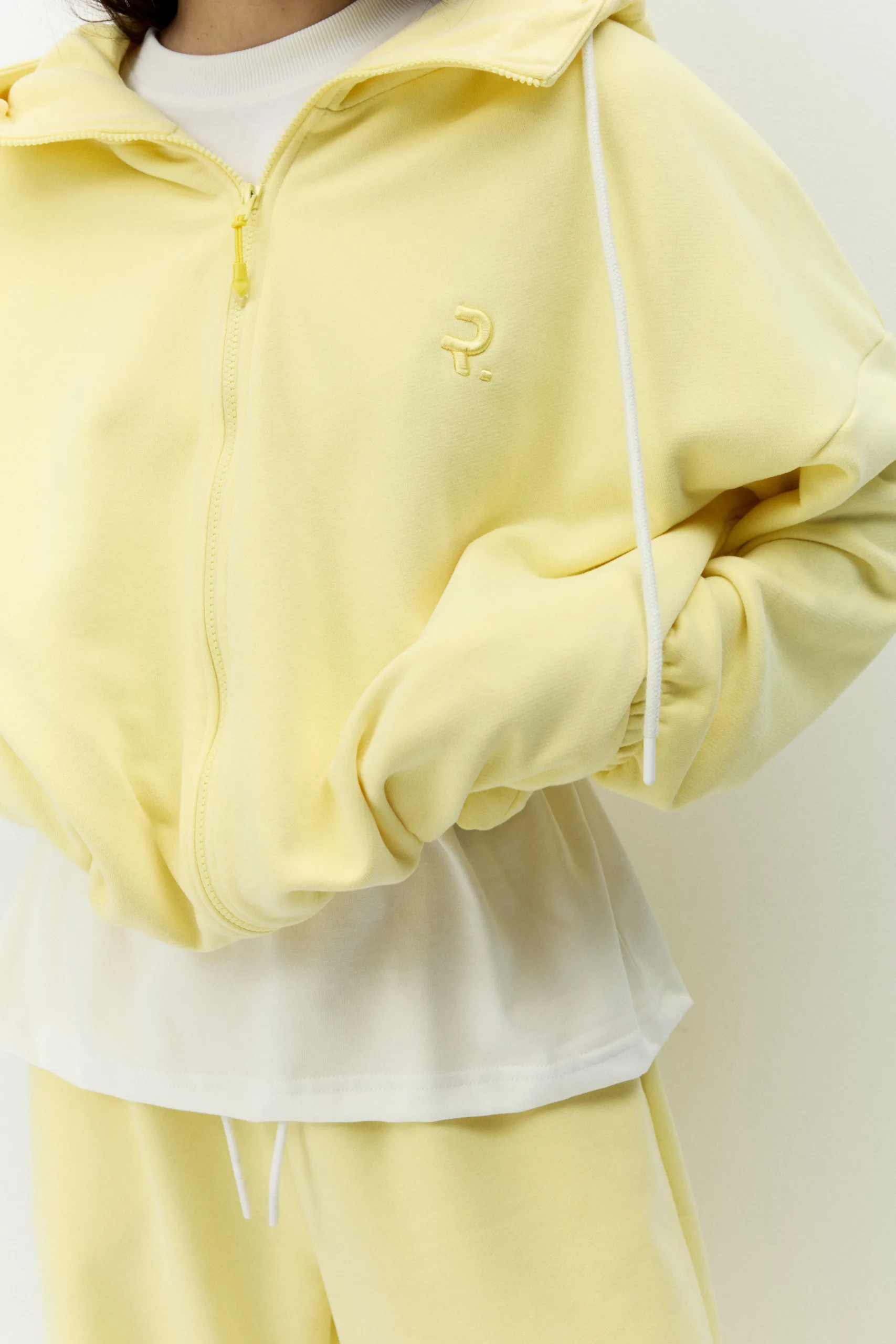 Жіноче oversized худі лимонного кольору з футеру діагональ преміум. Вільний силует, блискавка та м’яка фактура для casual образів. - 1C7A0588_Original Худі жіноче oversized лимонне на блискавці без утеплення - Close-up of a pale yellow zip-up hoodie with white drawstrings and a small embroidered logo on the left chest area.