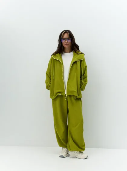 Person standing against a white wall in a lime-green tracksuit with a white undershirt and purple glasses, hands in pockets.