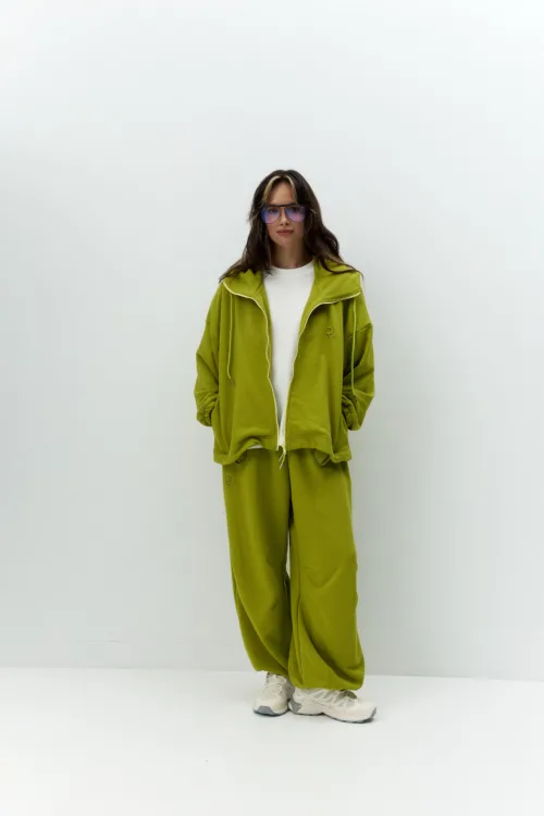 Full-body portrait of a person wearing an olive-green zip-up tracksuit over a white T-shirt, with blue-tinted sunglasses, standing against a plain white wall.