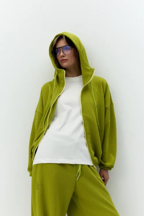 Woman wearing a lime-green hoodie and matching joggers with a white shirt, posing against a pale wall and blue-tinted sunglasses.